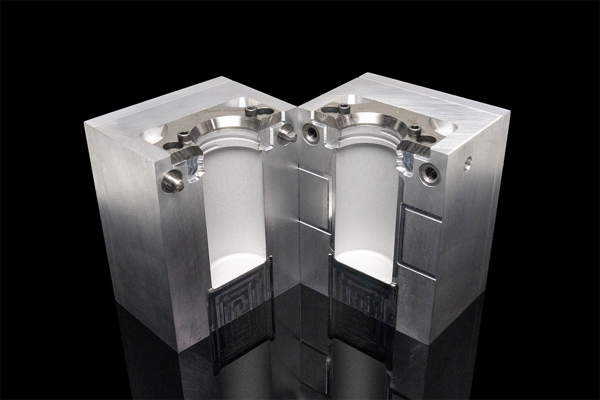 An image of a blow mold tool that was created with cnc manufacturing and metal 3d printing. The mold is a silver metal color on a black background, with mold halves open so you can see the inside cavity and neck inserts.