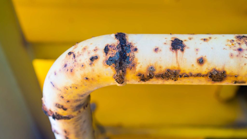 A metal pipe with a 90 degree bend shows spots of rusting on various parts of its surface, a result of pitting corrosion.