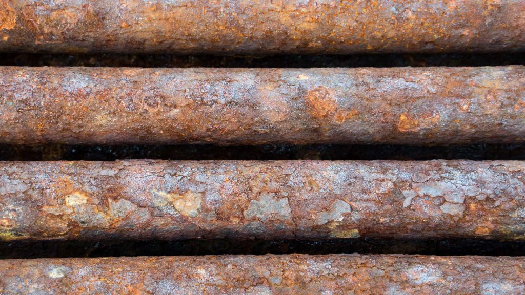 Several rusty pipes are stacked on top of or lay next to each other. The entire surface of each pipe is corroded.